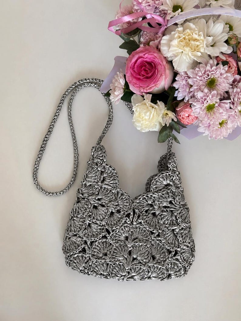 May include: A metallic silver crochet handbag featuring a repeating fan-like shell stitch pattern throughout the body. The bag includes a matching braided silver shoulder strap and an open top design, showcasing a textured, shiny finish.