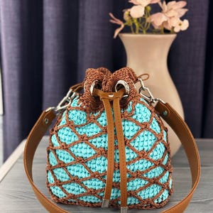Pode incluir: Uma bolsa de balde de croch&ecirc; feita &agrave; m&atilde;o com interior turquesa e rede marrom. A bolsa apresenta um fecho de cord&atilde;o marrom, uma al&ccedil;a de ombro de couro marrom e ferragens prateadas. Um acess&oacute;rio elegante e &uacute;nico.