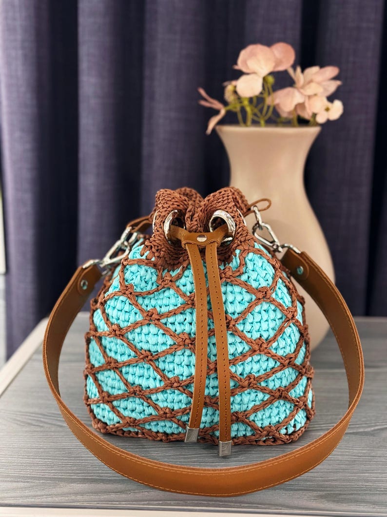 Pode incluir: Uma bolsa de balde de croch&ecirc; feita &agrave; m&atilde;o com interior turquesa e rede marrom. A bolsa apresenta um fecho de cord&atilde;o marrom, uma al&ccedil;a de ombro de couro marrom e ferragens prateadas. Um acess&oacute;rio elegante e &uacute;nico.