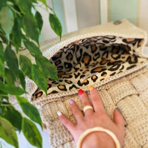 May include: A beige crocheted handbag featuring a woven texture and a flap closure. The interior is lined with a fabric displaying a brown, black, and tan leopard print pattern. A small rectangular white label is sewn onto the inner lining.