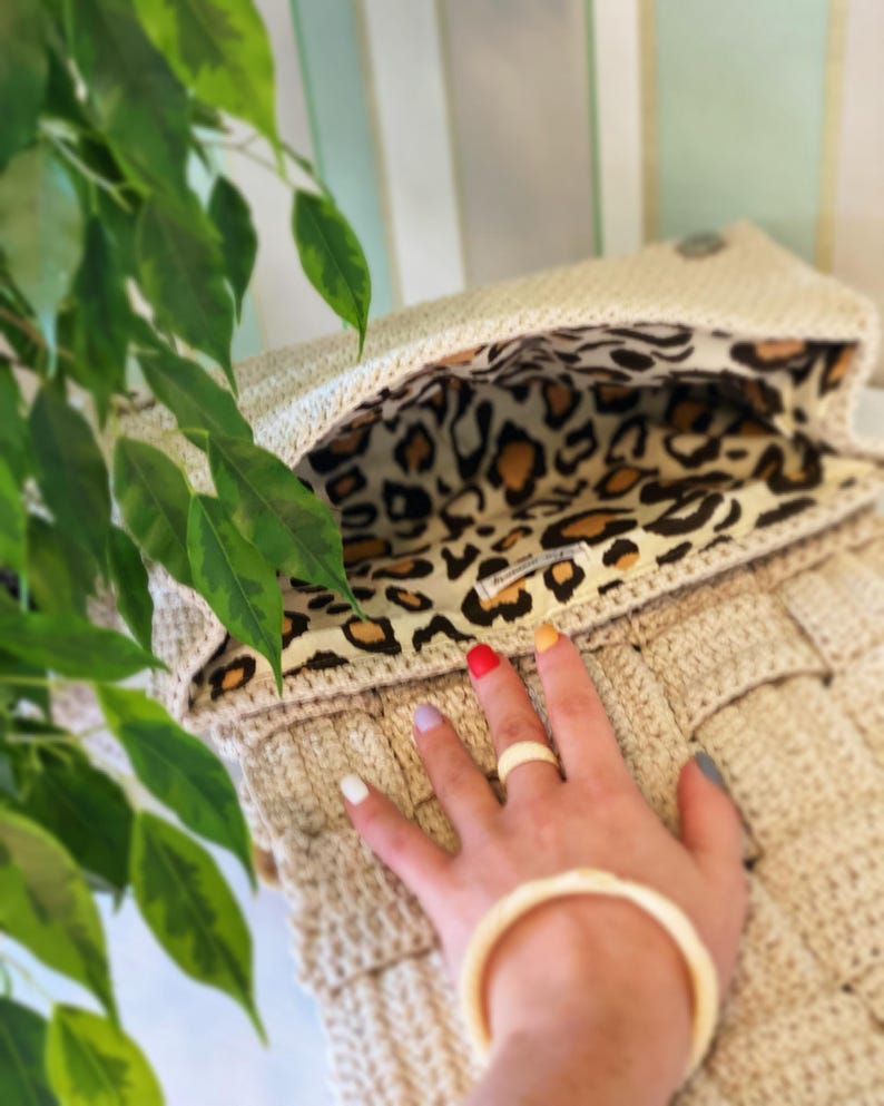 May include: A beige crocheted handbag featuring a woven texture and a flap closure. The interior is lined with a fabric displaying a brown, black, and tan leopard print pattern. A small rectangular white label is sewn onto the inner lining.