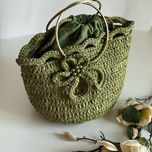 May include: A green woven handbag with a floral design and gold-colored handles. The bag has a drawstring closure and is lined with green fabric. The floral design is embellished with small, round beads. The bag is approximately 10 inches wide and 8 inches tall.