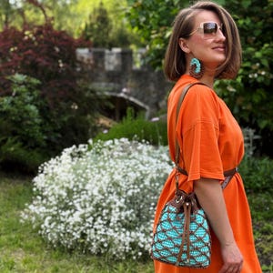 Pode incluir: Vestido laranja com cinto castanho e bolsa de croch&ecirc; turquesa e castanha com fecho de cord&atilde;o. A bolsa tem uma al&ccedil;a de couro castanho. A pessoa usa &oacute;culos de sol e brincos turquesa.