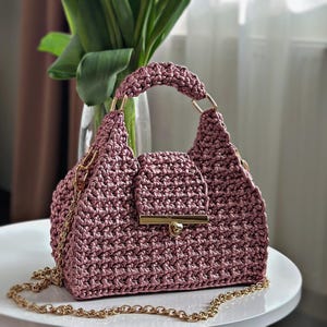 May include: A mauve-colored handbag crafted with a textured crochet pattern. The bag features a single top handle, a gold-toned metal turn-lock closure on the front flap, and a detachable gold-toned metal chain shoulder strap with lobster clasps.