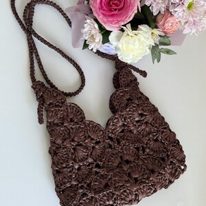 May include: A dark brown crochet handbag featuring a repeating shell stitch pattern and a matching braided shoulder strap. The bag has an open top design with knotted strap attachments on each side, showcasing a textured, handcrafted appearance.