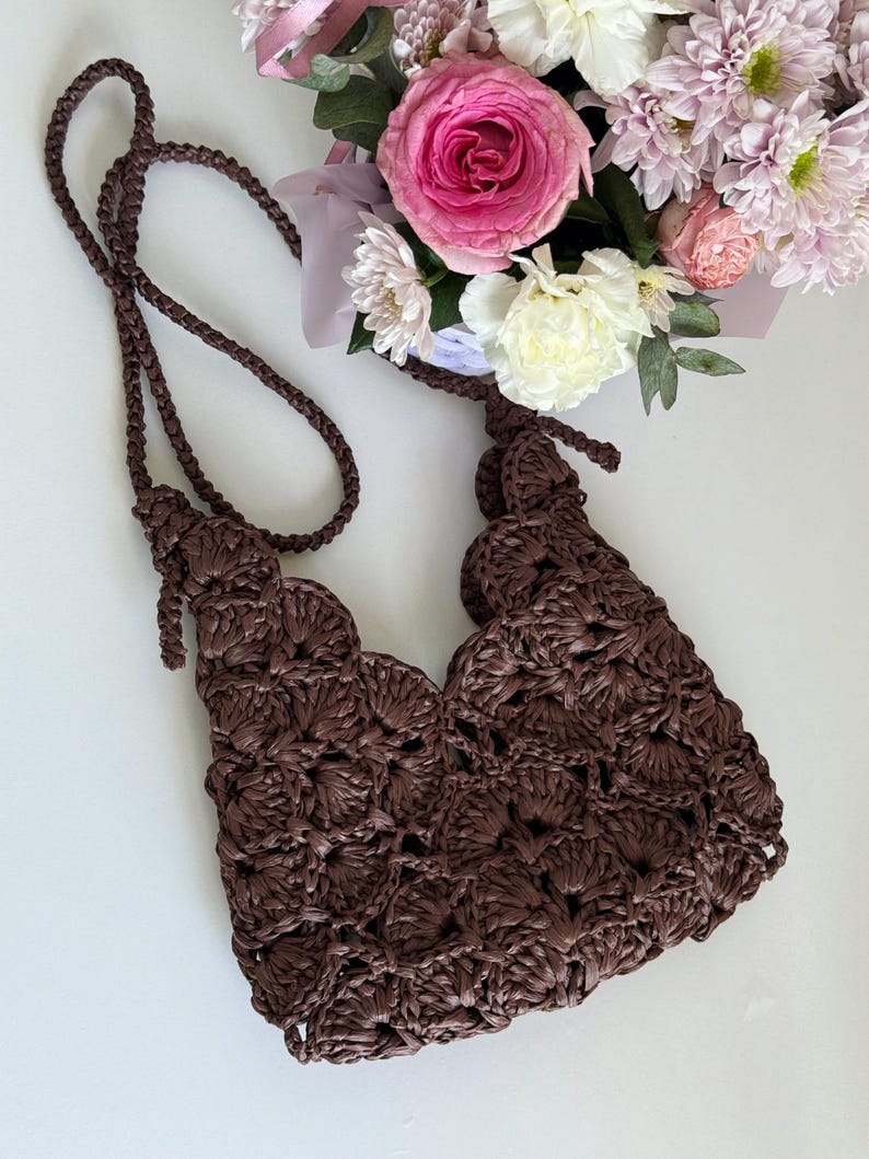 May include: A dark brown crochet handbag featuring a repeating shell stitch pattern and a matching braided shoulder strap. The bag has an open top design with knotted strap attachments on each side, showcasing a textured, handcrafted appearance.
