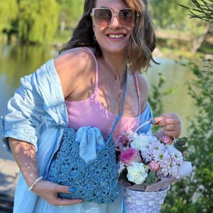 May include: A light blue crochet crossbody bag with a drawstring closure, featuring a textured shell pattern. The bag has a matching blue strap and is worn over a light blue button-down shirt and a pink ribbed tank top, paired with square sunglasses.