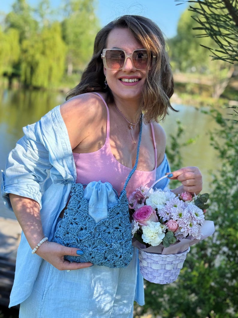 May include: A light blue crochet crossbody bag with a drawstring closure, featuring a textured shell pattern. The bag has a matching blue strap and is worn over a light blue button-down shirt and a pink ribbed tank top, paired with square sunglasses.