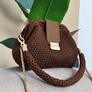 Puede incluir: Un bolso de mano de ganchillo marrón oscuro con estructura rígida, una solapa central de piel sintética lisa y un cierre de metal dorado. El bolso incluye un asa gruesa trenzada de color marrón y una correa de cadena dorada extraíble.
