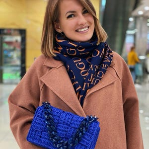 May include: A bright blue rectangular crochet handbag featuring a thick, chunky matching blue acrylic chain link strap. The bag has a woven texture and is paired with a navy blue scarf printed with repeating orange text in a bold, sans-serif font.