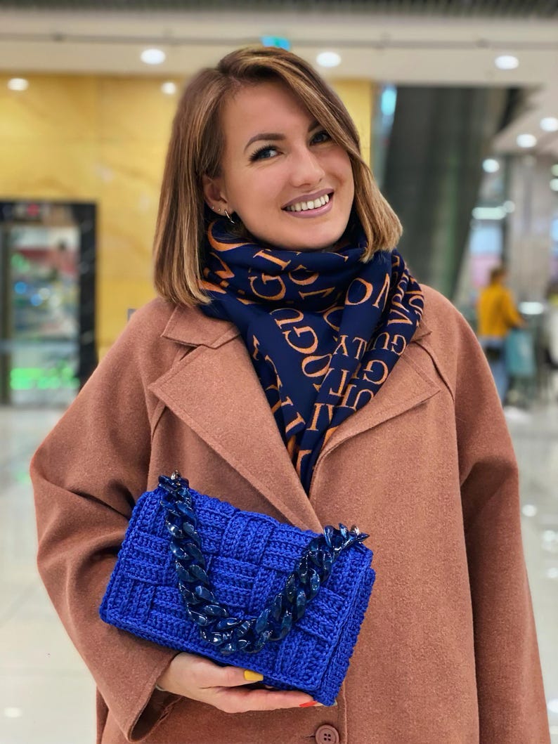 May include: A bright blue rectangular crochet handbag featuring a thick, chunky matching blue acrylic chain link strap. The bag has a woven texture and is paired with a navy blue scarf printed with repeating orange text in a bold, sans-serif font.