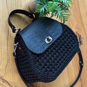 May include: Black handbag with a textured, woven body and a flap closure. The flap is made of black faux leather with a crocodile skin pattern. The bag has a silver clasp and a black strap. The bag is sitting on a wooden surface.