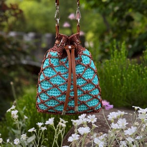 Pode incluir: Uma bolsa de balde de croch&ecirc; turquesa e marrom com fecho de cord&atilde;o de couro e al&ccedil;as de ombro. A bolsa apresenta um design de croch&ecirc; com padr&atilde;o de diamante e est&aacute; suspensa contra um fundo verde desfocado.