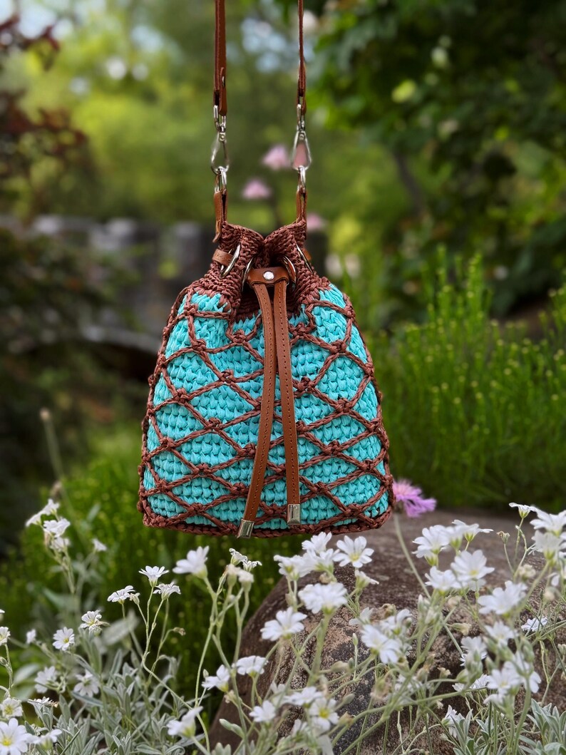Pode incluir: Uma bolsa de balde de croch&ecirc; turquesa e marrom com fecho de cord&atilde;o de couro e al&ccedil;as de ombro. A bolsa apresenta um design de croch&ecirc; com padr&atilde;o de diamante e est&aacute; suspensa contra um fundo verde desfocado.