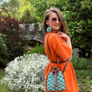 Pode incluir: Vestido laranja com uma bolsa de al&ccedil;a de couro marrom com um design de croch&ecirc; turquesa e marrom. A bolsa tem um fecho de cord&atilde;o. Brincos turquesa combinando s&atilde;o vis&iacute;veis. O fundo inclui vegeta&ccedil;&atilde;o e uma estrutura de pedra.