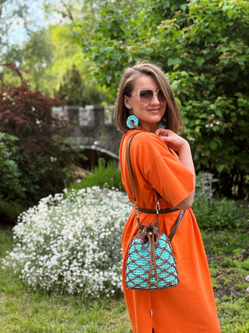 Pode incluir: Vestido laranja com uma bolsa de al&ccedil;a de couro marrom com um design de croch&ecirc; turquesa e marrom. A bolsa tem um fecho de cord&atilde;o. Brincos turquesa combinando s&atilde;o vis&iacute;veis. O fundo inclui vegeta&ccedil;&atilde;o e uma estrutura de pedra.