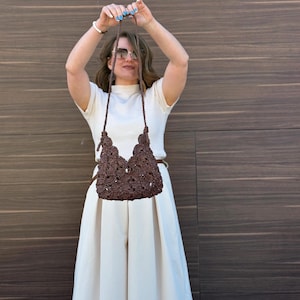 May include: A dark brown crochet handbag featuring a textured floral pattern and a thin matching shoulder strap. The bag has a scalloped top edge and is constructed from interconnected dark brown yarn elements in a repeating botanical design.