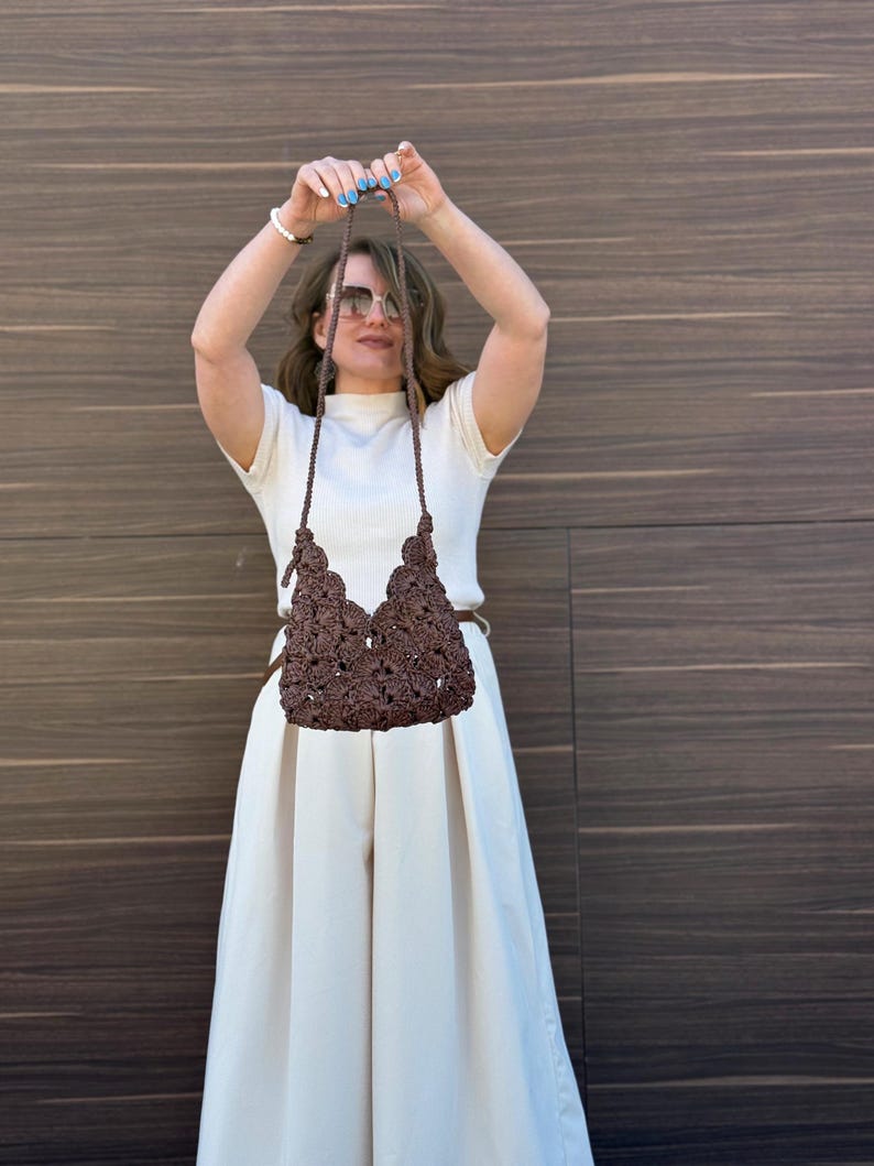 May include: A dark brown crochet handbag featuring a textured floral pattern and a thin matching shoulder strap. The bag has a scalloped top edge and is constructed from interconnected dark brown yarn elements in a repeating botanical design.