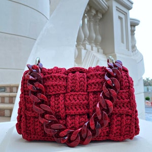 May include: A deep red, hand-crocheted handbag featuring a thick, woven texture and a chunky, translucent red acrylic chain link handle. The bag has a rectangular shape with silver-toned metal hardware connecting the handle to the main body.