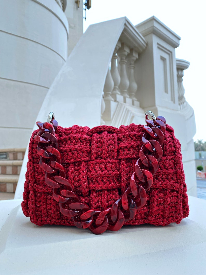 May include: A deep red, hand-crocheted handbag featuring a thick, woven texture and a chunky, translucent red acrylic chain link handle. The bag has a rectangular shape with silver-toned metal hardware connecting the handle to the main body.