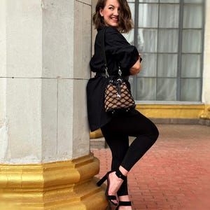 Pode incluir: Uma mulher vestindo um traje preto, incluindo uma camisa de manga comprida, leggings e sand&aacute;lias de salto alto. Ela est&aacute; carregando uma pequena bolsa estampada. O fundo apresenta uma coluna de pedra com detalhes dourados e uma janela.