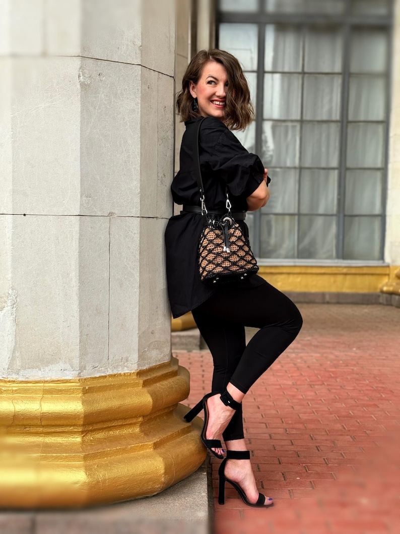 Pode incluir: Uma mulher vestindo um traje preto, incluindo uma camisa de manga comprida, leggings e sand&aacute;lias de salto alto. Ela est&aacute; carregando uma pequena bolsa estampada. O fundo apresenta uma coluna de pedra com detalhes dourados e uma janela.