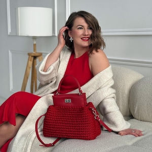 May include: A red crochet handbag with a matching red leather top handle and shoulder strap. The bag has a rectangular shape with a silver clasp. The bag is paired with a red dress and a cream-colored cardigan.