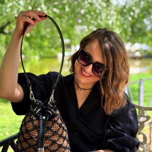 Pode incluir: Uma bolsa de balde de croch&ecirc; marrom e preta feita &agrave; m&atilde;o com uma al&ccedil;a de couro preta e fecho de cord&atilde;o. A bolsa tem um design exclusivo em forma de rede e ferragens prateadas. A bolsa &eacute; segurada por uma pessoa.