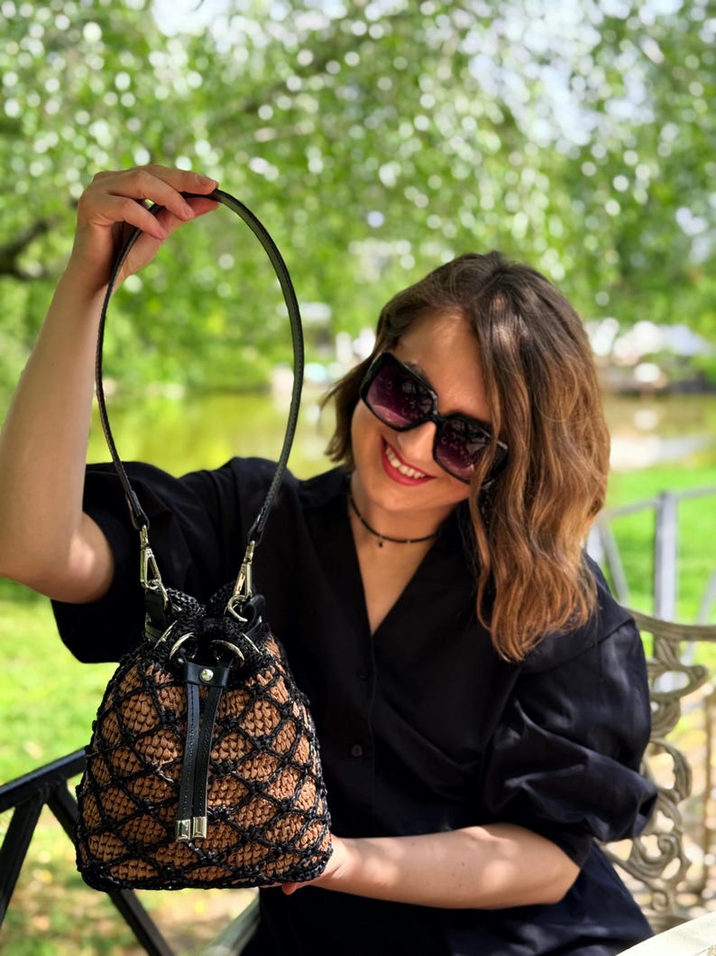 Pode incluir: Uma bolsa de balde de croch&ecirc; marrom e preta feita &agrave; m&atilde;o com uma al&ccedil;a de couro preta e fecho de cord&atilde;o. A bolsa tem um design exclusivo em forma de rede e ferragens prateadas. A bolsa &eacute; segurada por uma pessoa.