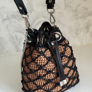 Pode incluir: Uma bolsa de balde tecida com uma sobreposi&ccedil;&atilde;o de padr&atilde;o de diamante preto. A bolsa apresenta um interior castanho, al&ccedil;as de couro preto e ferragens prateadas. Uma al&ccedil;a de corrente e um fecho de cord&atilde;o tamb&eacute;m s&atilde;o vis&iacute;veis.