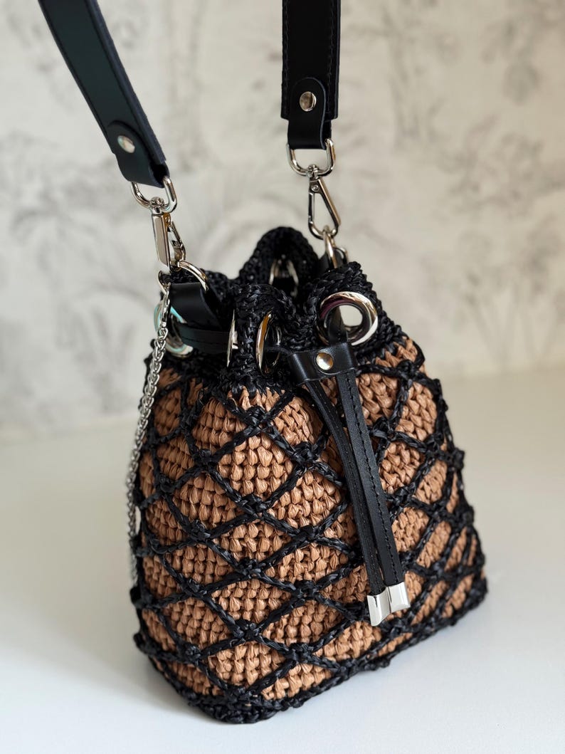 Pode incluir: Uma bolsa de balde tecida com uma sobreposi&ccedil;&atilde;o de padr&atilde;o de diamante preto. A bolsa apresenta um interior castanho, al&ccedil;as de couro preto e ferragens prateadas. Uma al&ccedil;a de corrente e um fecho de cord&atilde;o tamb&eacute;m s&atilde;o vis&iacute;veis.