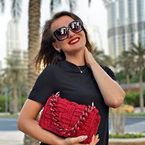 May include: A rectangular, deep red crocheted handbag featuring a thick, chunky, matching red acrylic chain link handle. The bag has a woven, basket-like texture and is held in front of a plain black short-sleeved shirt.