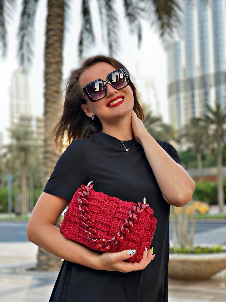 May include: A rectangular, deep red crocheted handbag featuring a thick, chunky, matching red acrylic chain link handle. The bag has a woven, basket-like texture and is held in front of a plain black short-sleeved shirt.