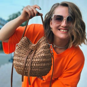 Puede incluir: Un bolso de cubo marrón tejido con cierre de cordón y correas de cuero. El bolso tiene una correa para el hombro y un cierre plateado. El bolso lo sostiene una persona que lleva una camisa naranja.