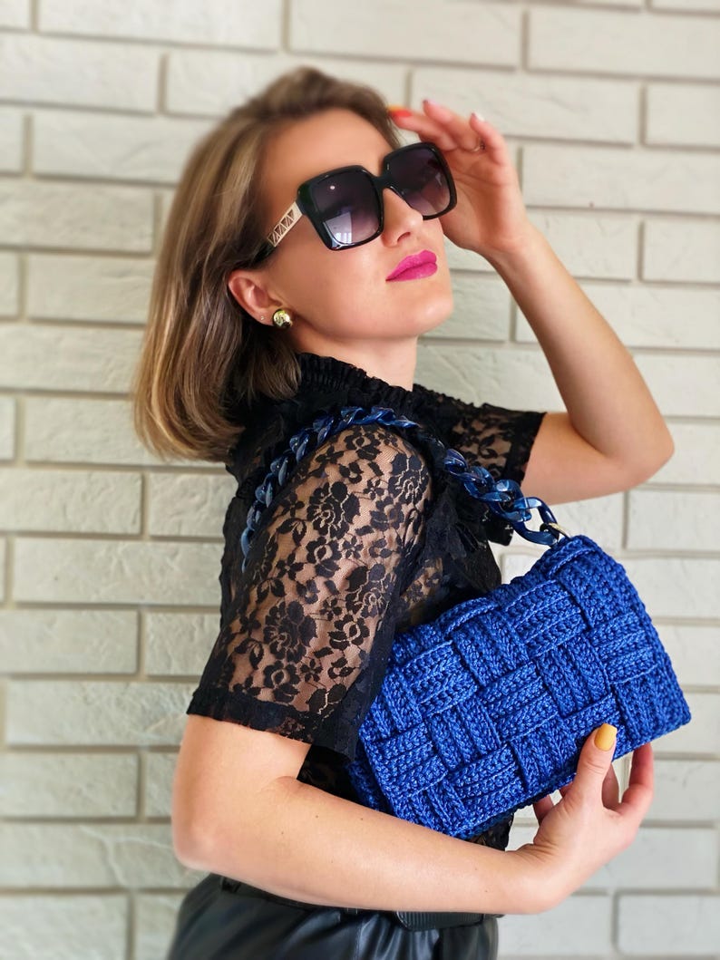 May include: A vibrant royal blue woven handbag featuring a thick, chunky matching blue plastic chain link handle. The bag has a distinct square-patterned texture created by interlaced cord material, providing a structured and tactile appearance.