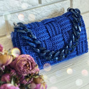 May include: A rectangular handbag crafted from royal blue yarn in a woven basket-weave pattern. The bag features a thick, chunky dark blue acrylic chain link handle attached to silver-toned metal rings on each side of the top edge.