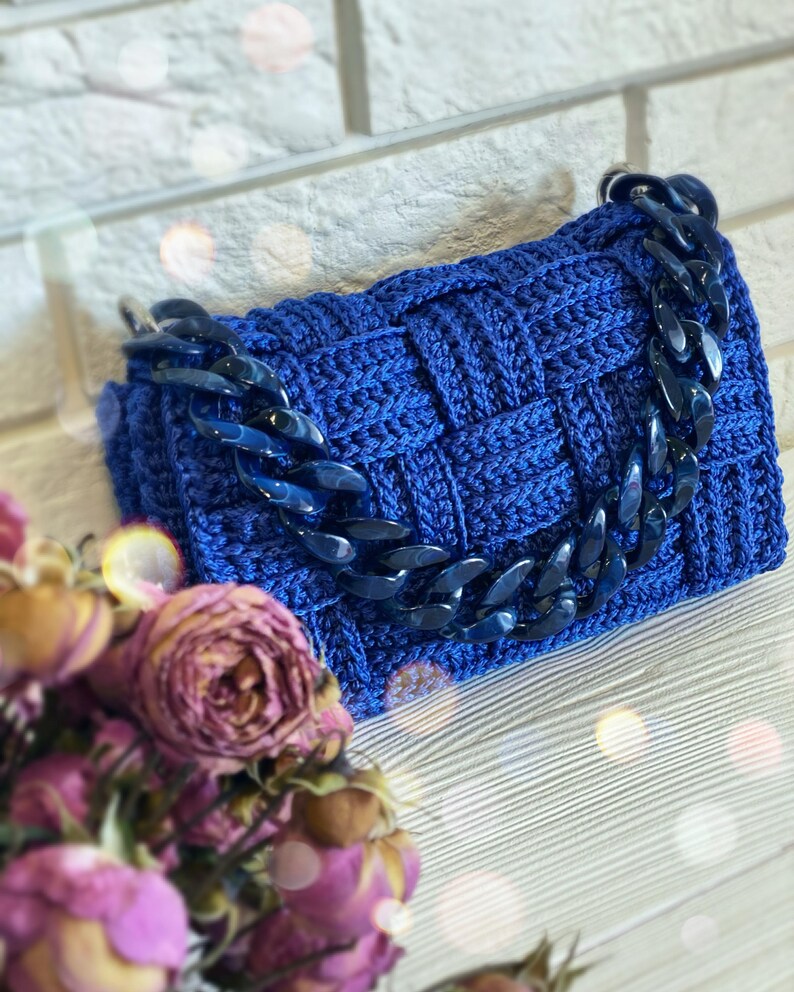 May include: A rectangular handbag crafted from royal blue yarn in a woven basket-weave pattern. The bag features a thick, chunky dark blue acrylic chain link handle attached to silver-toned metal rings on each side of the top edge.