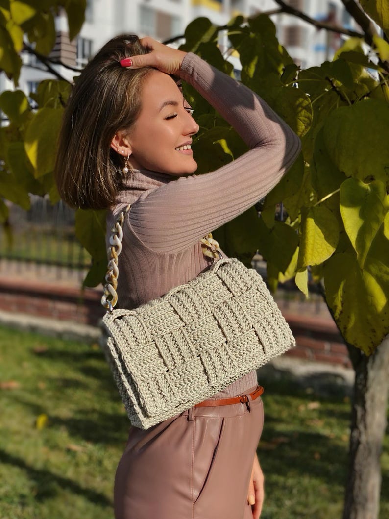 May include: A rectangular crochet handbag in a light beige color featuring a woven basket-weave pattern and a thick, chunky chain-link handle made of marbled cream and tan acrylic links, attached to the bag with silver-toned metal hardware.