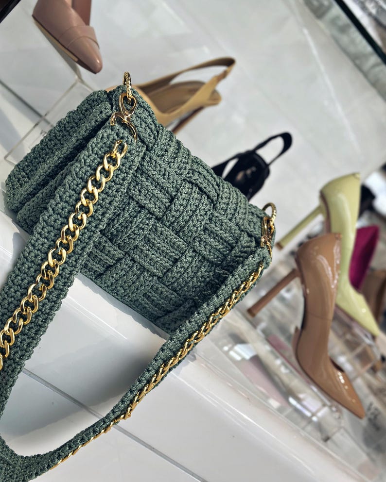 May include: A sage green crocheted handbag featuring a woven texture and a gold-tone metal chain strap. The bag has a rectangular shape with a flap closure and is constructed from thick yarn, showcasing a detailed interlocking pattern throughout.