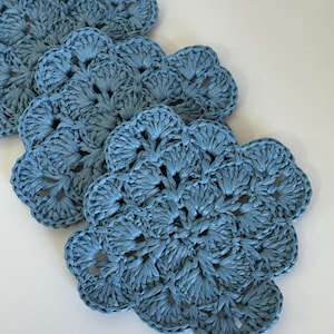 May include: A set of four dusty blue crochet coasters, each featuring a circular, flower-like pattern with scalloped edges. The coasters are crafted from a textured, fibrous material and are arranged in a slightly overlapping diagonal line.