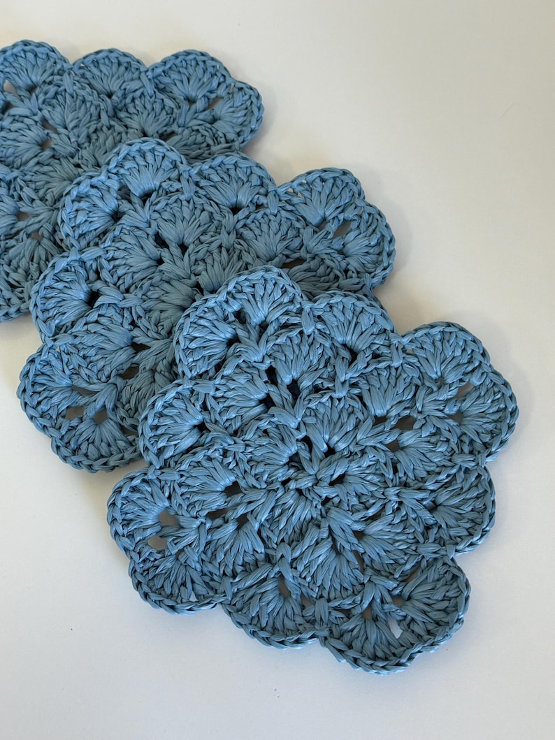 May include: A set of four dusty blue crochet coasters, each featuring a circular, flower-like pattern with scalloped edges. The coasters are crafted from a textured, fibrous material and are arranged in a slightly overlapping diagonal line.