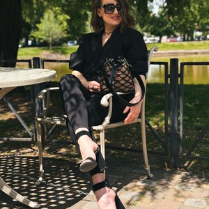 Pode incluir: Uma pessoa com um traje preto, incluindo um blazer, cal&ccedil;as e sand&aacute;lias de salto alto, est&aacute; sentada numa cadeira ao ar livre. Ela est&aacute; usando &oacute;culos de sol e segurando uma pequena bolsa preta e bege. O fundo apresenta um parque com &aacute;rvores e um lago.