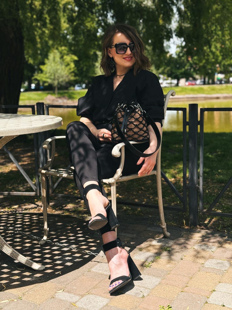 Pode incluir: Uma pessoa com um traje preto, incluindo um blazer, cal&ccedil;as e sand&aacute;lias de salto alto, est&aacute; sentada numa cadeira ao ar livre. Ela est&aacute; usando &oacute;culos de sol e segurando uma pequena bolsa preta e bege. O fundo apresenta um parque com &aacute;rvores e um lago.
