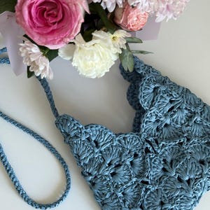 May include: A dusty blue crochet handbag featuring a scalloped edge pattern and a braided shoulder strap. The bag is positioned next to a bouquet of pink and white roses and chrysanthemums, showcasing the intricate texture of the yarn work.