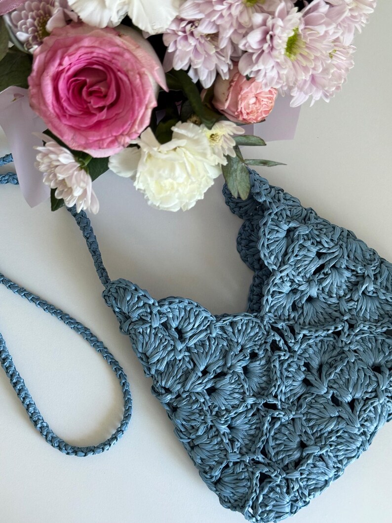 May include: A dusty blue crochet handbag featuring a scalloped edge pattern and a braided shoulder strap. The bag is positioned next to a bouquet of pink and white roses and chrysanthemums, showcasing the intricate texture of the yarn work.
