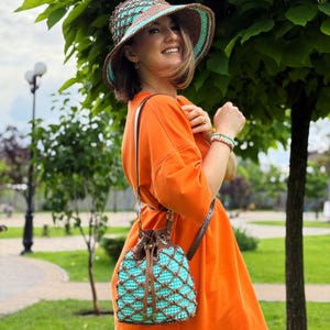 Pode incluir: Um chap&eacute;u de croch&ecirc; e uma bolsa com cord&atilde;o a condizer em tons de turquesa, castanho e creme. O chap&eacute;u tem uma aba larga e a bolsa apresenta um padr&atilde;o de diamante. A bolsa tem uma al&ccedil;a de couro castanho. A mulher est&aacute; vestida com um vestido laranja.