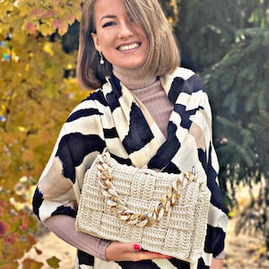 May include: A beige woven handbag featuring a thick, chunky, marbled acrylic chain strap in shades of brown and cream. The bag has a rectangular, structured shape with a grid-like texture and a flap closure, crafted from a light tan cord material.