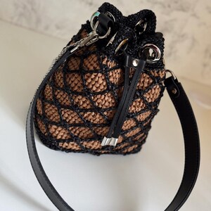 Pode incluir: Uma bolsa de balde tecida em preto e bege com uma al&ccedil;a de couro preta. A bolsa apresenta um fecho de cord&atilde;o e ferragens prateadas. O padr&atilde;o tecido cria um efeito de treli&ccedil;a e a bolsa tem uma forma estruturada.