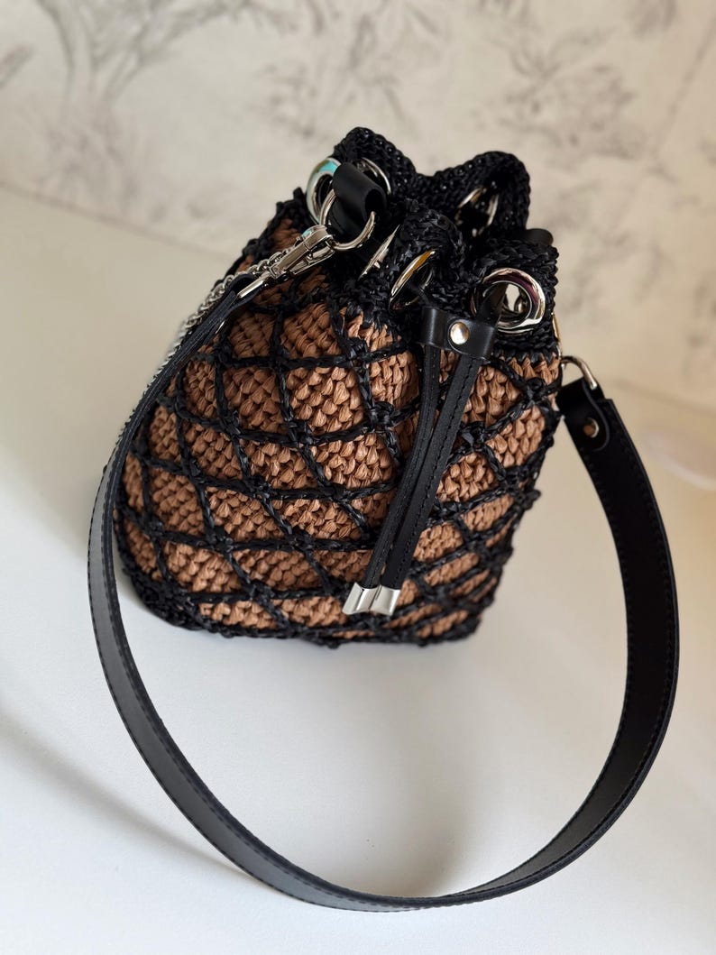 Pode incluir: Uma bolsa de balde tecida em preto e bege com uma al&ccedil;a de couro preta. A bolsa apresenta um fecho de cord&atilde;o e ferragens prateadas. O padr&atilde;o tecido cria um efeito de treli&ccedil;a e a bolsa tem uma forma estruturada.