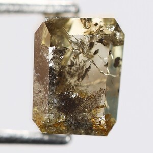 May include: A light brown, rectangular, faceted gemstone with inclusions of black and gold. The gemstone is held by a pair of tweezers.